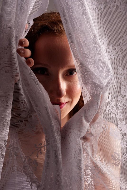 kobieta patrząca zza firany w pięknym wieczorowym makijażu - sesja zdjęciowa